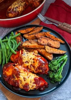 a black plate topped with chicken, green beans and sweet potato wedges next to a red casserole dish