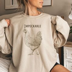 "This funny chicken laying an egg \"Impeckable\" (impeccable) sweatshirt is a cute gift for crazy chicken lovers whether they are living in the country, in cities, on farms, or in the suburbs. Makes a great gift for yourself and others. Unisex sizing. Order up for an oversized look, plus sizes available. DESCRIPTION * This unisex heavy blend crewneck sweatshirt is pure comfort.  * The collar is ribbed knit, so it retains its shape even after washing. There are no itchy side seams. * 50% pre-shrunk cotton, 50% polyester. This combination helps designs come out looking fresh and beautiful.  * Fabric weight: 8.0 oz/yd² (271.25 g/m²) * Air-jet spun yarn with a soft feel and reduced pilling. Quarter-turned body to avoid crease down the middle. * 1 × 1 athletic rib-knit cuffs and waistband with Chicken Sweatshirt, Country Mom Aesthetic, Chicken Mom, Crazy Chicken Lady, Chicken Shirts, Chicken Lady, Trending Today, Selling Clothes, Mom Shirts