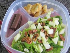 a plastic container filled with salad and nuts on top of a table next to a piece of chocolate