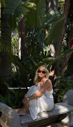 a woman sitting on top of a stone bench in front of palm trees and greenery