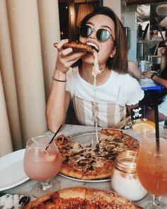 a woman sitting at a table with pizza and drinks in front of her eating the pizza