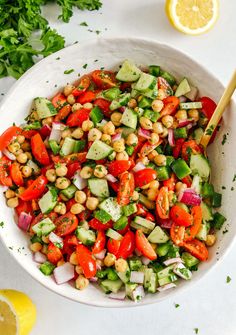 a white bowl filled with cucumber, tomato and chickpea salad next to lemon wedges