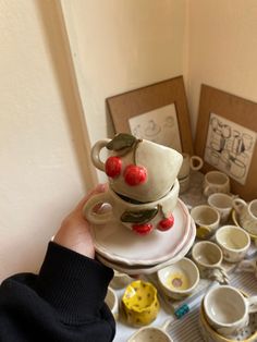 a person is holding a teapot with cherries on it in front of cups and saucers