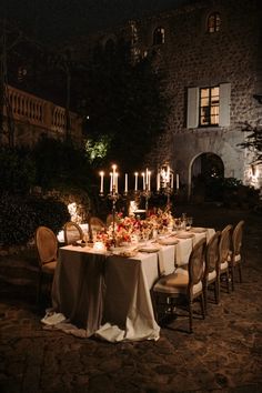 an outdoor dinner table set up with candles and flowers in front of a stone building