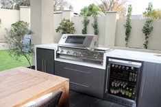 an outdoor kitchen with stainless steel appliances and table in the back yard, surrounded by greenery