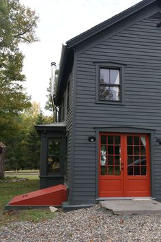 a gray house with red door and windows on gravel area next to trees, grass and rocks