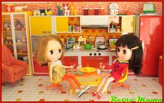 two dolls sitting at a table in a dollhouse kitchen with orange walls and floor tiles