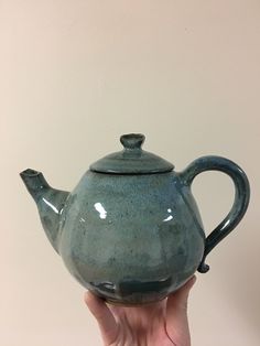 a hand holding a green teapot in front of a white wall with the lid open