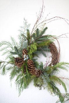 a wreath with pine cones and greenery hanging on the wall next to a white wall