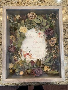 a frame with flowers and greenery in it on top of a marble countertop