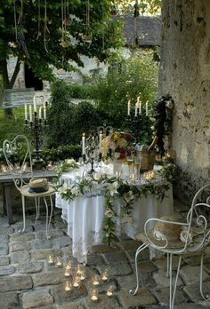 an outdoor dining table with candles and flowers on it, surrounded by greenery in the background