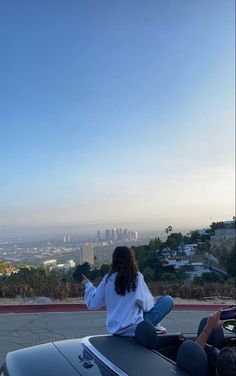 a woman sitting on top of a convertible looking out at the city from atop a hill