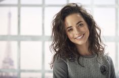 a woman with long hair smiling at the camera in front of a wall and window