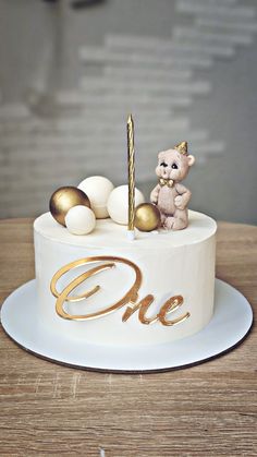 a white cake with gold decorations and a small teddy bear on top that says one