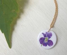 "This is my original design of a tiny, sweet violet flower. I am fascinated by violets; their deep jewel-like intense colors, delicate petals, lush heart-shaped leaves, and spindly stems. A big patch of violets comes up in our backyard every spring and when they bloom, my older daughter picks a handful for me to put in my small glass vase on the windowsill by the kitchen sink. They are the brightest color in the room! This is one of my smallest pendants; it measures 5/8\" across, smaller than a Handmade Delicate Purple Necklace, Purple Flower Charm Necklace As A Gift, Purple Flower Necklace With Charm As A Gift, Purple Flower Necklace With Flower Charm, Purple Flower Charm Necklace, Purple Flower Pendant With Pressed Flowers, Purple Flower Pendant Necklace, Purple Flower Shaped Necklace With Pressed Flowers, Purple Flower Pendant Necklace For Birth Flower