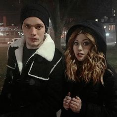 a man and woman standing next to each other on a sidewalk at night with street lights in the background