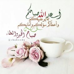 there is a coffee cup and some pink flowers on the table with arabic writing above it