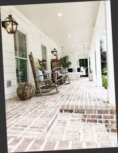 two rocking chairs on the front porch of a house with white siding and brick flooring