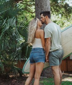 a man and woman kissing in front of a tree with a surfboard behind them