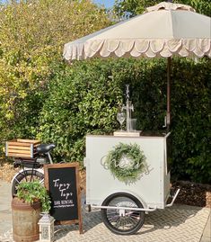 an ice cream cart with a white umbrella and some plants on the sidewalk next to it