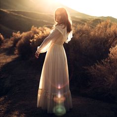 a woman in a long white dress standing on a hill with the sun behind her