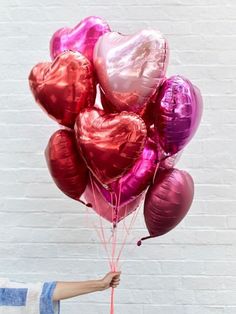 a person holding some heart shaped balloons in front of a white brick wall with pink and red foil