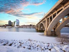 a bridge over water with snow on the ground and buildings in the background at sunset