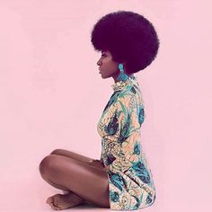 a woman with an afro sitting on the ground in front of a pink background and looking off to the side