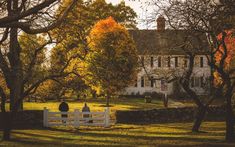 two people sitting on a white fence in front of a large house surrounded by trees