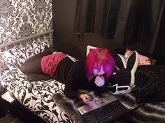 a woman with pink hair laying on a bed using a laptop