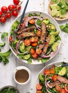 two white bowls filled with steak and vegetables next to tomatoes, cucumbers, lettuce, and avocado