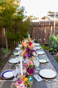 a long wooden table with plates and flowers on it is set for an outdoor dinner