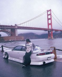 a man sitting on the hood of a white sports car in front of a bridge