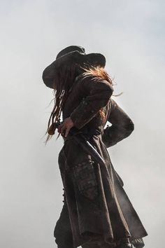 a man in a long coat and hat standing on top of a hill with his back to the camera