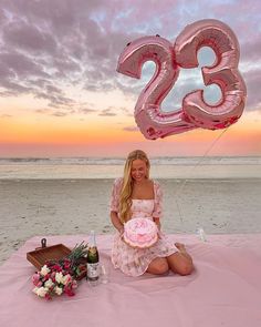 a woman sitting on the beach with a cake and balloons in front of her at sunset