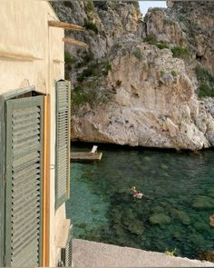 two people are swimming in the water near some rocks and buildings with shutters on them