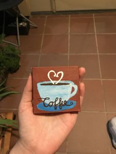 a hand holding up a small coaster with a cup of coffee painted on the front