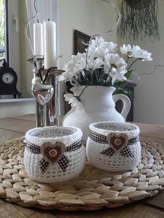 two white crocheted mugs sitting on top of a table next to a vase with flowers