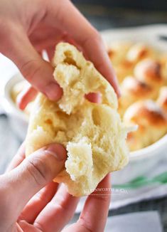 two hands holding a piece of bread in front of a casserole dish filled with rolls