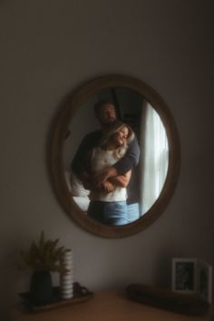 a man holding a woman in front of a round mirror on the wall next to a table