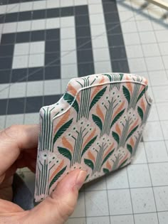 a hand holding an orange and green patterned wallet on a tiled floor next to a black and white checkered wall