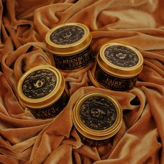 three small jars sitting on top of a brown cloth next to some gold colored material