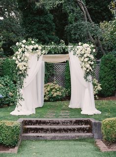 an outdoor wedding ceremony with white flowers and draping on the arch, surrounded by greenery