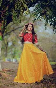 a woman in a yellow skirt and red top standing under a tree with her hands on her head