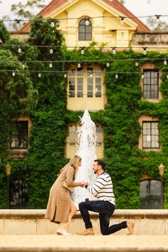 a man kneeling down next to a woman in front of a fountain with greenery