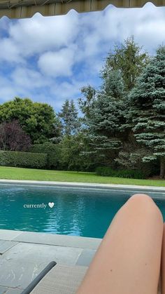 a woman's legs sitting next to a swimming pool with trees in the background