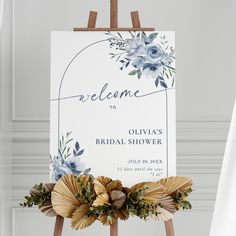 a welcome sign with flowers and greenery sits on an easel in front of a white wall