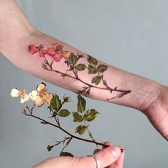 a woman's arm with flowers on it and leaves in the foreground, while another person holds out their hand