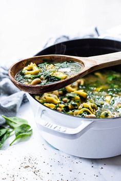 a wooden spoon in a white pot filled with pasta and spinach soup on a marble surface