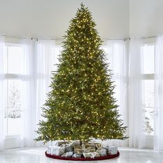 a christmas tree with presents under it in a living room next to two large windows